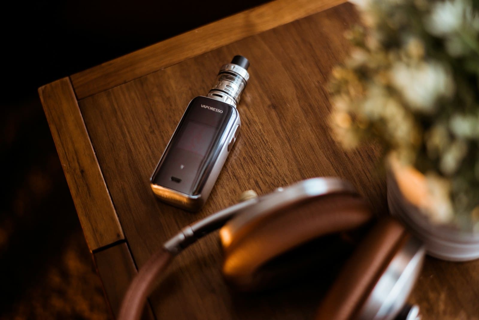 A compact silver pod vape kit resting on a wooden table next to a pair of headphones