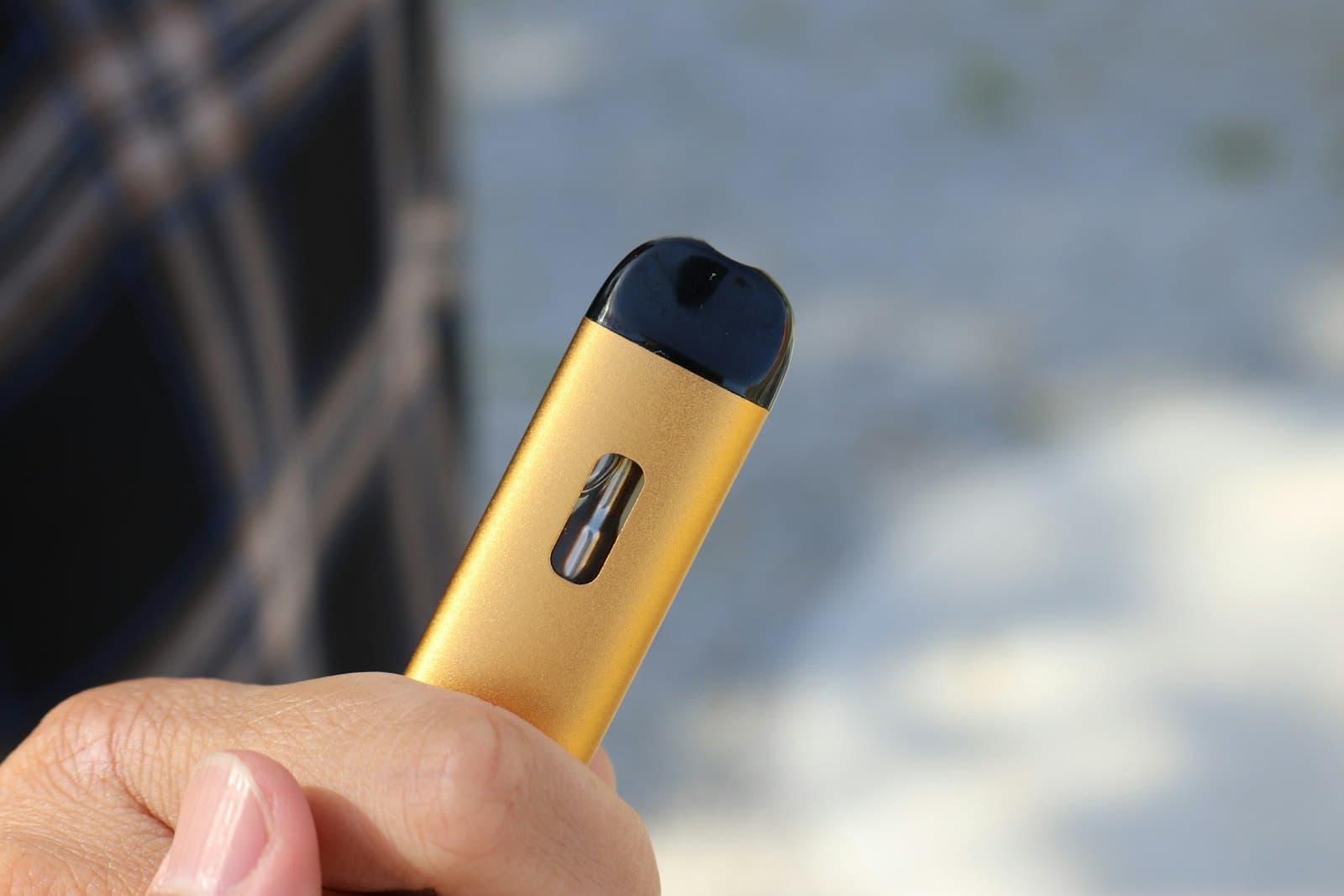 A slim, gold-coloured draw-activated pod vape device held between two fingers outdoors